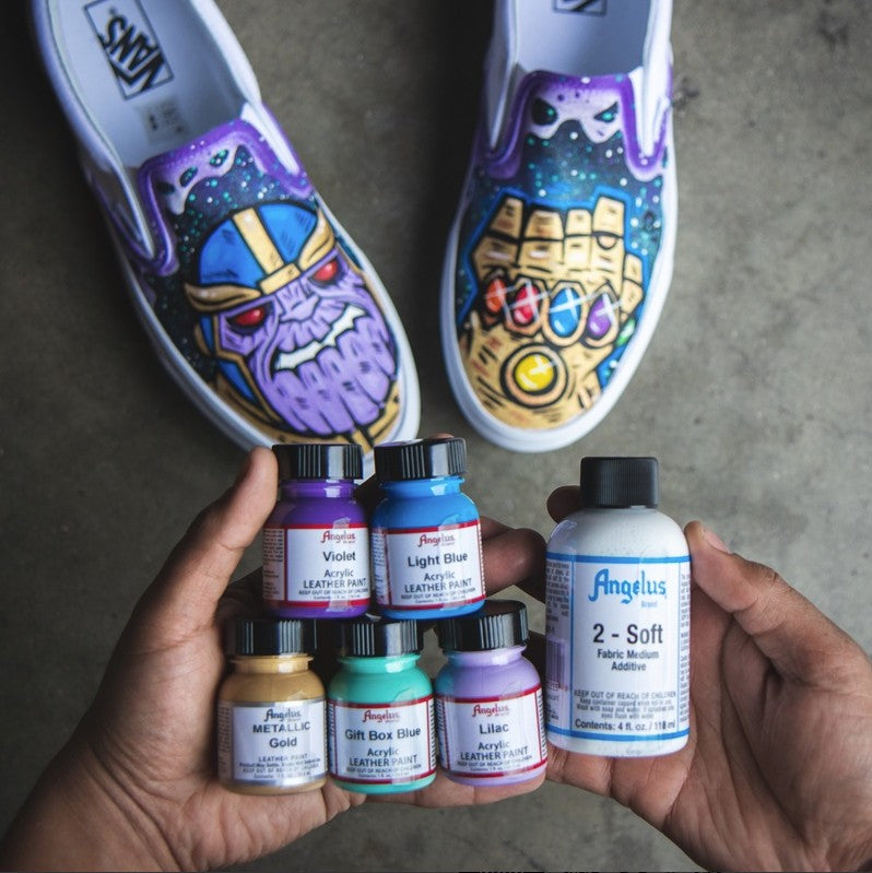 2 perechi de vans customizati cu thanos si infinity gauntlet, 5 sticle de vopsea acrilica angelus pentru piele de 29ml, violet, albastru deschis, gold, gift box blue si lilac si aditivul 2-soft angelus de 118ml tinute in maini pe un fundal de ciment