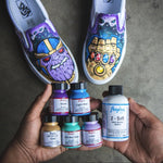 2 perechi de vans customizati cu thanos si infinity gauntlet, 5 sticle de vopsea acrilica angelus pentru piele de 29ml, violet, albastru deschis, gold, gift box blue si lilac si aditivul 2-soft angelus de 118ml tinute in maini pe un fundal de ciment